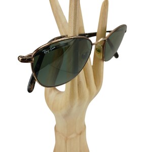 May include: Gold-toned Ray-Ban sunglasses with green lenses. The sunglasses are displayed on a wooden hand.