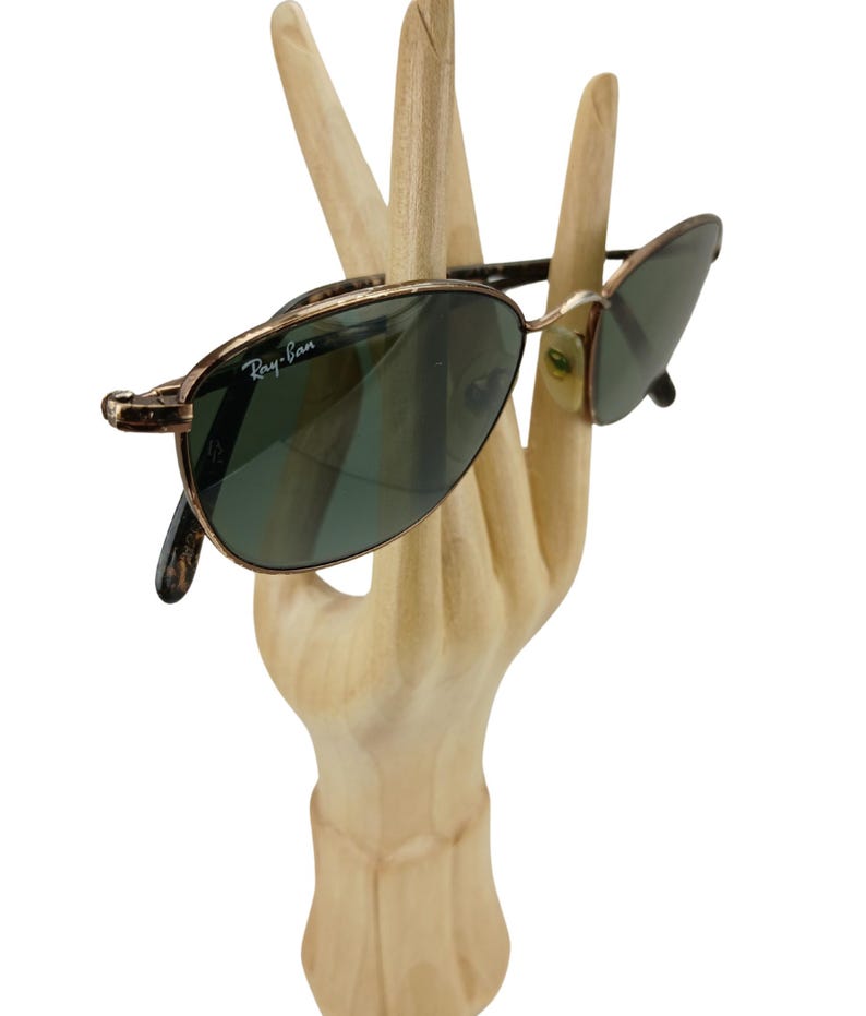 May include: Gold-toned Ray-Ban sunglasses with green lenses. The sunglasses are displayed on a wooden hand.