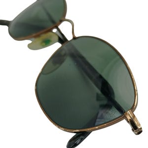 May include: A pair of gold-rimmed sunglasses with green lenses. The sunglasses are in focus and the background is blurred.
