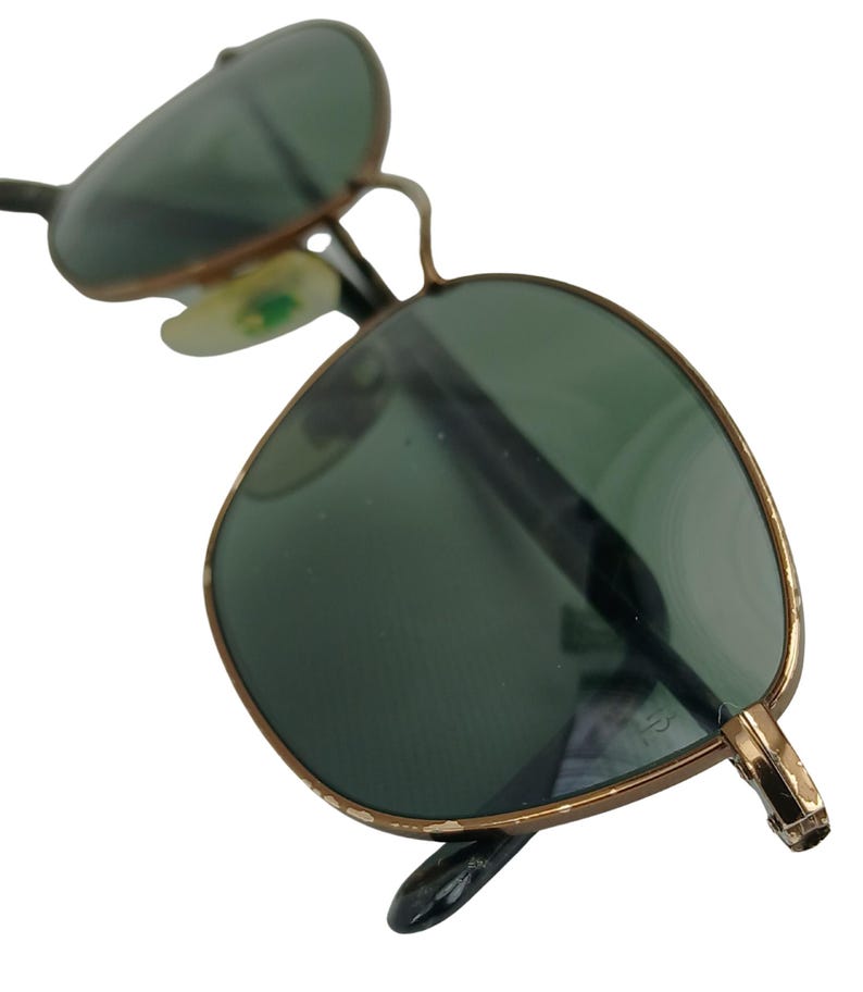 May include: A pair of gold-rimmed sunglasses with green lenses. The sunglasses are in focus and the background is blurred.