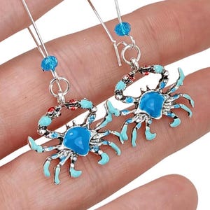 May include: A pair of silver dangle earrings featuring blue and red enamel crab charms with turquoise blue cabochon accents. The earrings have a silver hook closure and a blue crystal bead on each earring.