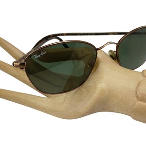 May include: A pair of brown metal Ray-Ban sunglasses with green lenses. The sunglasses are on a wooden hand.