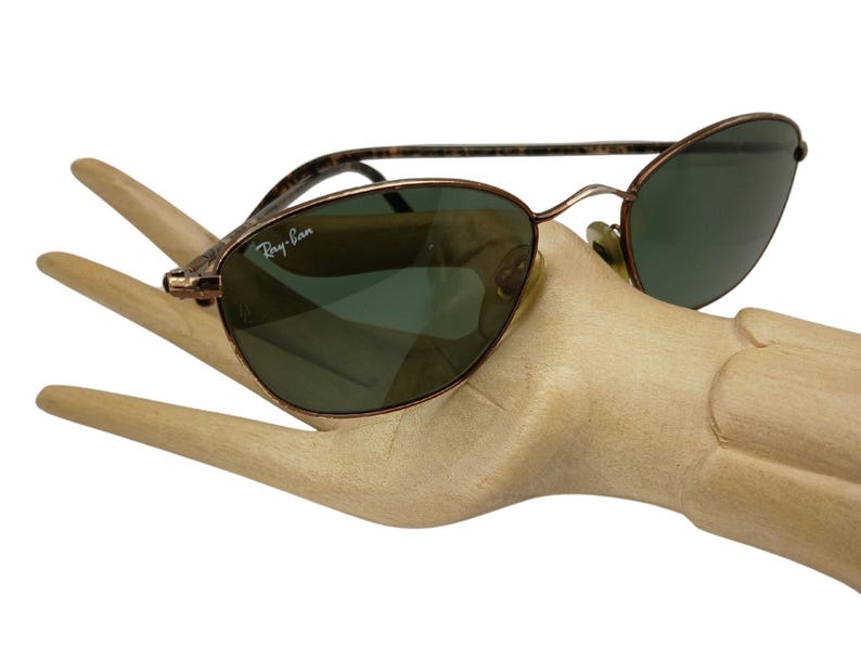 May include: A pair of brown metal Ray-Ban sunglasses with green lenses. The sunglasses are on a wooden hand.