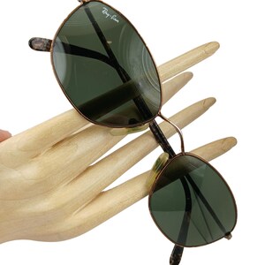 May include: Vintage Ray-Ban sunglasses with a gold metal frame and green lenses. The sunglasses have a rounded, oval shape.