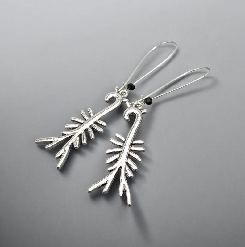 May include: A pair of silver-toned earrings with a unique, stylized design. Each earring features a long, curved hook and a decorative element resembling a stylized plant or feather. Small black beads accent the top of each earring.