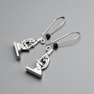 May include: A pair of silver-toned microscope earrings with black bead accents. The earrings feature a detailed miniature microscope design, suspended from long, curved ear wires. The background is a solid gray.