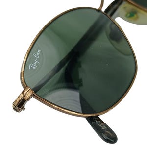May include: Gold-tone Ray-Ban sunglasses with green lenses. The sunglasses have a metal frame and the Ray-Ban logo is visible on the left lens.
