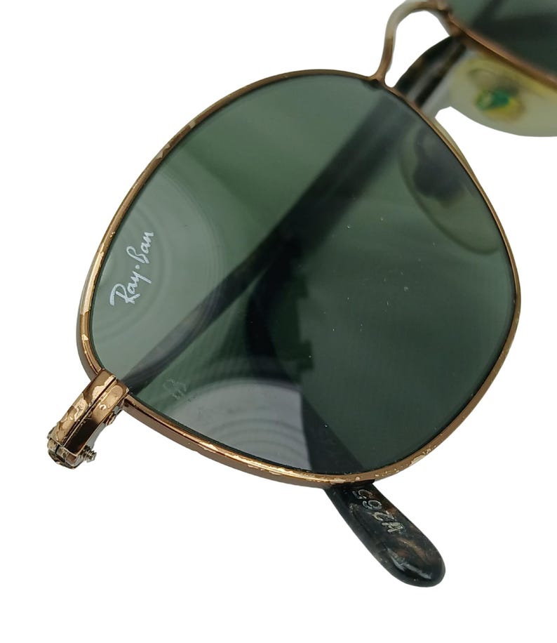 May include: Gold-tone Ray-Ban sunglasses with green lenses. The sunglasses have a metal frame and the Ray-Ban logo is visible on the left lens.