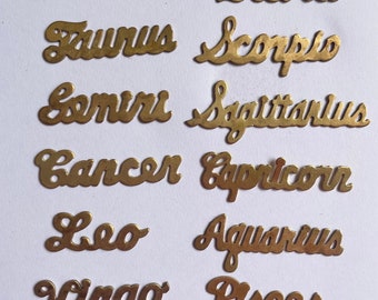May include: Twelve gold-coloured metal zodiac signs, each with a different astrological symbol. The signs are arranged in two rows of six, with the names of the signs written in a cursive font.