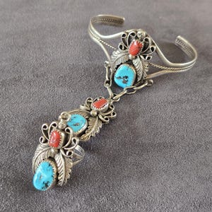 Pode incluir: Uma pulseira escrava de prata composta por um bracelete ligado por uma corrente a um anel. A joia é decorada com várias pedras ovais de turquesa e coral vermelho cravadas em metal ornamentado com padrões de folhas e detalhes em volutas.