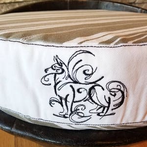 May include: A white and gray striped dog bed with a black embroidered design of a dog with a curly tail. The bed is round and has a black wooden base.