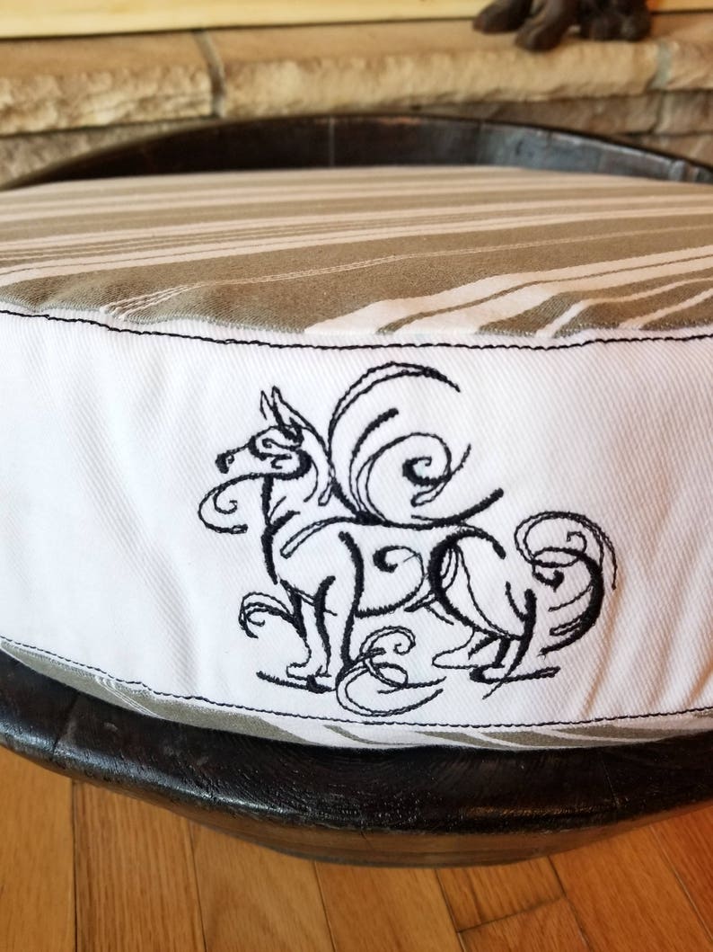 May include: A white and gray striped dog bed with a black embroidered design of a dog with a curly tail. The bed is round and has a black wooden base.
