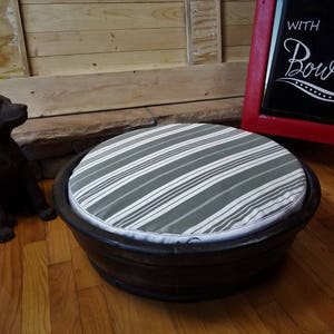 May include: A round pet bed made from a wooden barrel with a gray and white striped cushion. The bed is sitting on a wooden floor. The text "With My Bowl" is written on a red chalkboard sign in the background.
