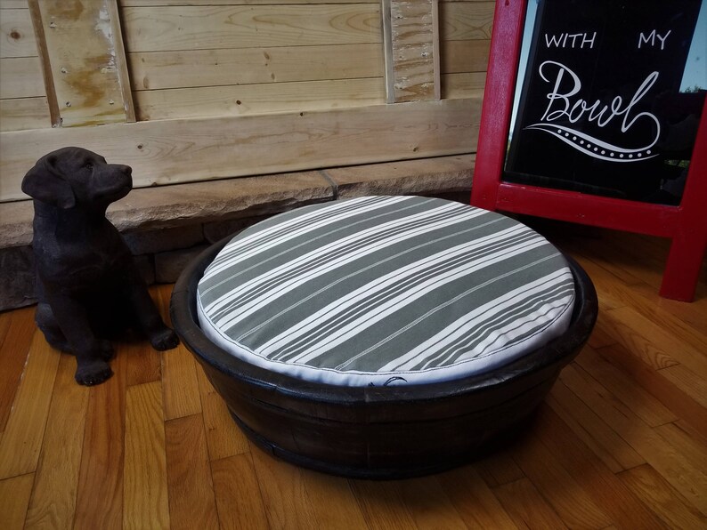 May include: A round pet bed made from a wooden barrel with a gray and white striped cushion. The bed is sitting on a wooden floor. The text "With My Bowl" is written on a red chalkboard sign in the background.