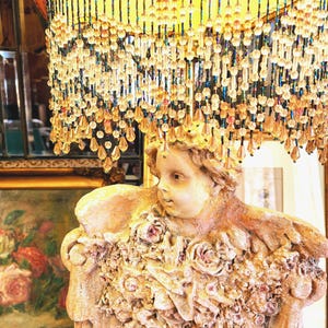Puede incluir: Una l&aacute;mpara vintage con una pantalla decorativa de cuentas y una base de estatua. La pantalla es naranja con un ribete granate y cuentas colgantes. La estatua es beige y presenta un querub&iacute;n con detalles florales.