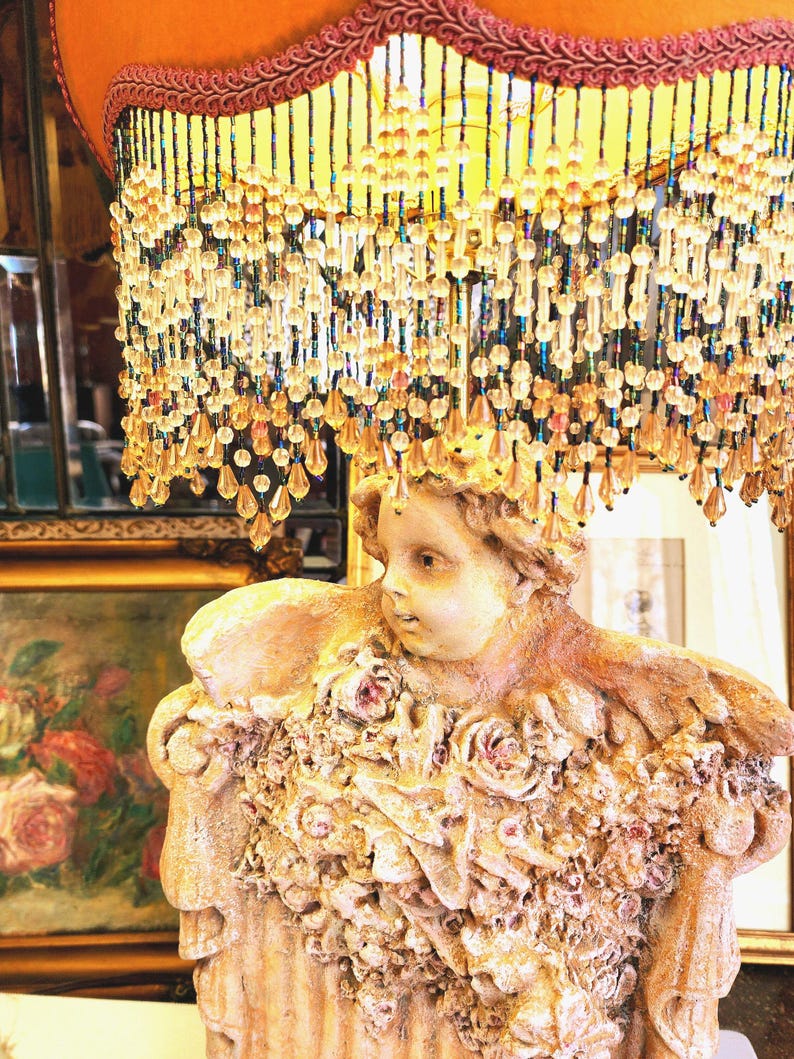 Puede incluir: Una l&aacute;mpara vintage con una pantalla decorativa de cuentas y una base de estatua. La pantalla es naranja con un ribete granate y cuentas colgantes. La estatua es beige y presenta un querub&iacute;n con detalles florales.