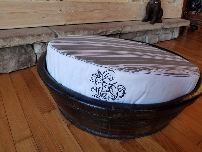 May include: A round, black, wooden pet bed with a white and gray striped cushion. The cushion has a black embroidered design of a dog with a floral pattern.