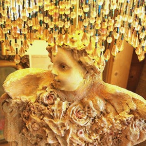 Puede incluir: Una l&aacute;mpara decorativa con una figura esculpida y detalles florales. La figura es de color beige claro, con un rostro detallado y una superficie texturizada. La l&aacute;mpara est&aacute; adornada con cuentas colgantes en tonos dorados y &aacute;mbar.