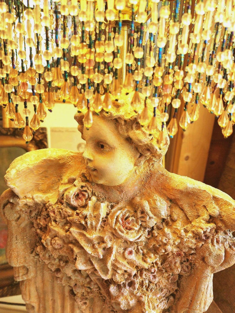 Puede incluir: Una l&aacute;mpara decorativa con una figura esculpida y detalles florales. La figura es de color beige claro, con un rostro detallado y una superficie texturizada. La l&aacute;mpara est&aacute; adornada con cuentas colgantes en tonos dorados y &aacute;mbar.