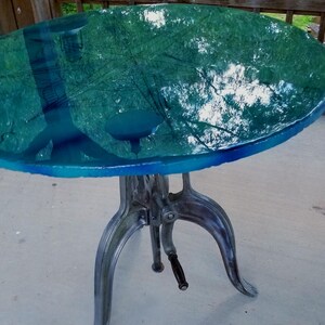 May include: A round, blue-green table with a clear epoxy finish. The table has a cast iron base with three legs.