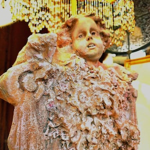 Puede incluir: Estatua antigua detallada de la cara y la parte superior del cuerpo de un ni&ntilde;o, con un dise&ntilde;o texturizado similar a flores. La estatua es principalmente marr&oacute;n con detalles en blanco y rosa. Un candelabro decorativo con cuentas de cristal cuelga encima.