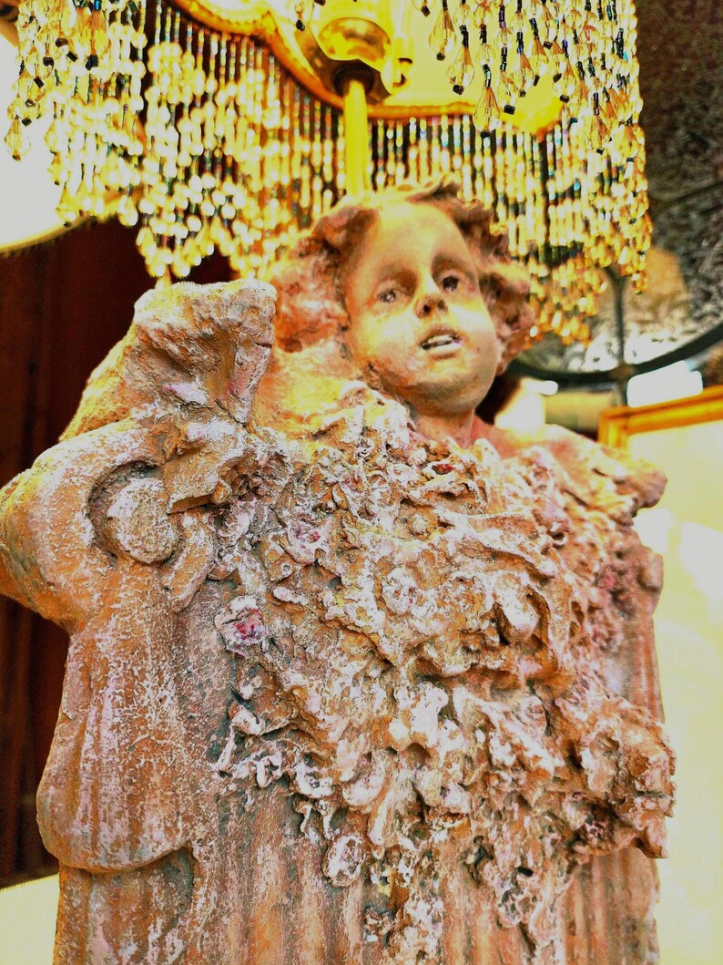 Puede incluir: Estatua antigua detallada de la cara y la parte superior del cuerpo de un ni&ntilde;o, con un dise&ntilde;o texturizado similar a flores. La estatua es principalmente marr&oacute;n con detalles en blanco y rosa. Un candelabro decorativo con cuentas de cristal cuelga encima.