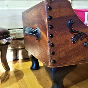 May include: A wooden dog bed with black leather accents and a red and white striped cushion. The bed has black metal legs and is decorated with a black and gold floral design.