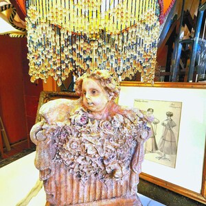 Puede incluir: L&aacute;mpara ornamentada con base de estatua de querub&iacute;n, adornada con detalles florales. La pantalla naranja tiene flecos de cuentas y adornos decorativos. Una ilustraci&oacute;n vintage enmarcada est&aacute; en el fondo.