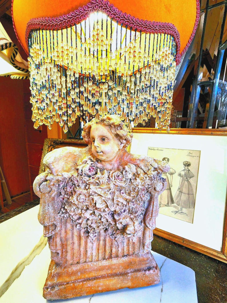 Puede incluir: L&aacute;mpara ornamentada con base de estatua de querub&iacute;n, adornada con detalles florales. La pantalla naranja tiene flecos de cuentas y adornos decorativos. Una ilustraci&oacute;n vintage enmarcada est&aacute; en el fondo.