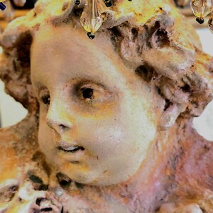 Puede incluir: Primer plano de una escultura antigua y desgastada de un rostro de querub&iacute;n. La escultura es una mezcla de tonos beige, bronce y rosa, con una superficie texturizada. Encima de la cabeza, adornos de cristal con detalles dorados y azules.