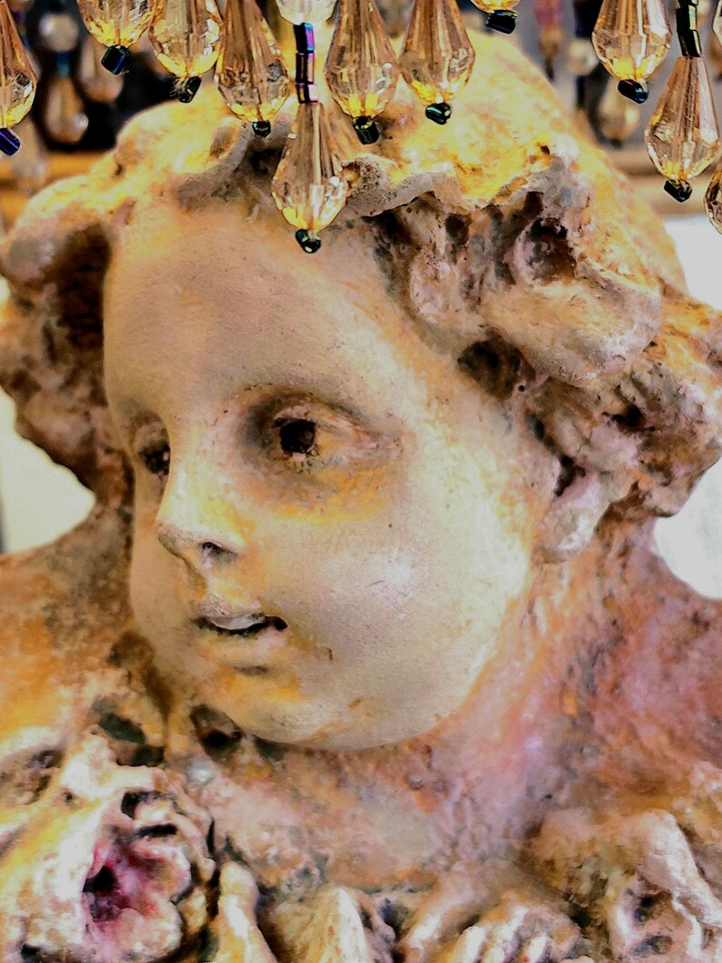 Puede incluir: Primer plano de una escultura antigua y desgastada de un rostro de querub&iacute;n. La escultura es una mezcla de tonos beige, bronce y rosa, con una superficie texturizada. Encima de la cabeza, adornos de cristal con detalles dorados y azules.