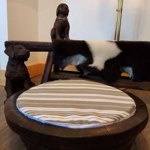 May include: A round, wooden pet bed with a striped cushion. The bed is made from a repurposed barrel and has a natural wood finish. The cushion is white and gray with horizontal stripes.