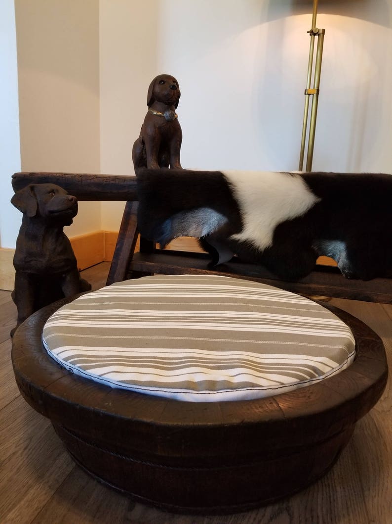 May include: A round, wooden pet bed with a striped cushion. The bed is made from a repurposed barrel and has a natural wood finish. The cushion is white and gray with horizontal stripes.