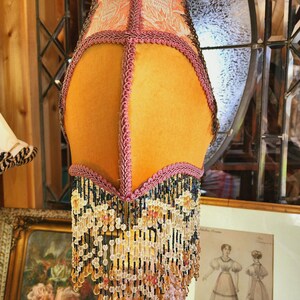 Puede incluir: Una pantalla de l&aacute;mpara vintage con un dise&ntilde;o floral melocot&oacute;n y amarillo. La pantalla de la l&aacute;mpara est&aacute; adornada con flecos de cuentas rosas y negras. La l&aacute;mpara es una pieza decorativa con un dise&ntilde;o &uacute;nico y ornamentado.