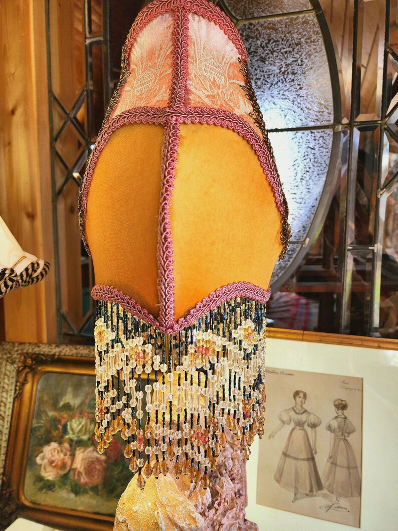 Puede incluir: Una pantalla de l&aacute;mpara vintage con un dise&ntilde;o floral melocot&oacute;n y amarillo. La pantalla de la l&aacute;mpara est&aacute; adornada con flecos de cuentas rosas y negras. La l&aacute;mpara es una pieza decorativa con un dise&ntilde;o &uacute;nico y ornamentado.