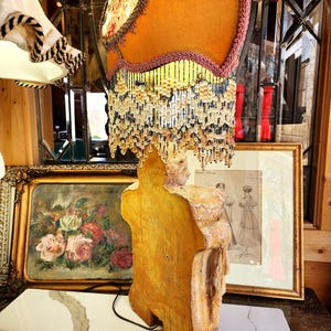 Puede incluir: Una l&aacute;mpara de mesa de estilo vintage con una pantalla de color amarillo mostaza, adornada con un estampado floral y flecos de cuentas. La base de la l&aacute;mpara es una figura tallada en tonos dorados. El fondo incluye obras de arte enmarcadas y una pared espejada.