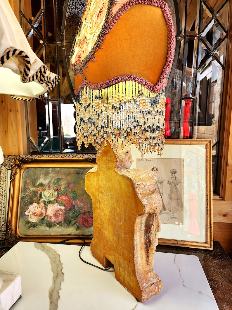 Puede incluir: Una l&aacute;mpara de mesa de estilo vintage con una pantalla de color amarillo mostaza, adornada con un estampado floral y flecos de cuentas. La base de la l&aacute;mpara es una figura tallada en tonos dorados. El fondo incluye obras de arte enmarcadas y una pared espejada.