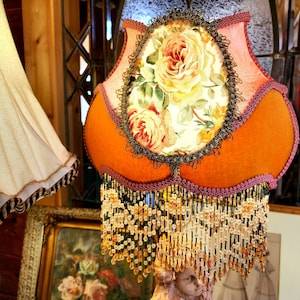 Puede incluir: Una pantalla de l&aacute;mpara naranja con un dise&ntilde;o floral, con un panel ovalado central con rosas. La pantalla tiene un fleco de cuentas y est&aacute; adornada con detalles en rosa y negro. La l&aacute;mpara es de estilo vintage.