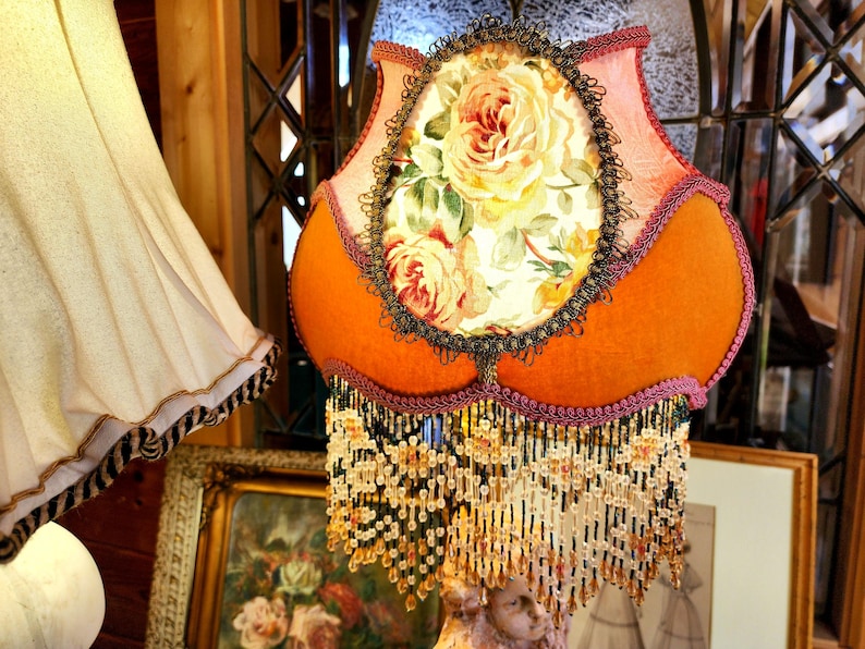 Puede incluir: Una pantalla de l&aacute;mpara naranja con un dise&ntilde;o floral, con un panel ovalado central con rosas. La pantalla tiene un fleco de cuentas y est&aacute; adornada con detalles en rosa y negro. La l&aacute;mpara es de estilo vintage.