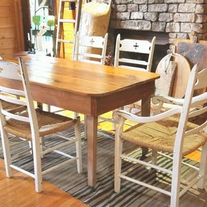 May include: A rustic wooden table with a light brown finish, surrounded by six white chairs with woven seats. The chairs have a simple design with a cross-shaped detail on the back. The table is set in a room with wood paneling and a brick fireplace.