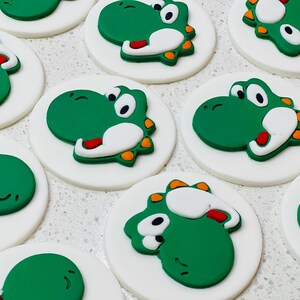 May include: A selection of Yoshi-themed fondant cupcake toppers. Each topper features a green Yoshi character with white, red, and orange details, set on a white circular base. The toppers are arranged on a light-coloured surface.