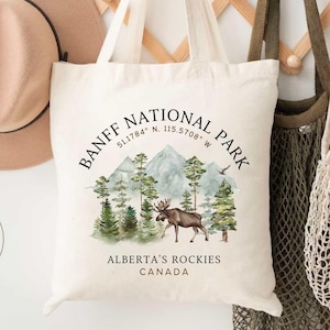 May include: A beige tote bag with the text "Banff National Park" and an illustration of a moose and mountains. A tan hat and two other bags hang on a wooden rack. The text "Alberta's Rockies Canada" is also printed on the bag.