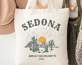 May include: A white canvas tote bag with the text "SEDONA" in black lettering. The bag features a graphic of a desert landscape with a sunset, mountains, and cacti. The text "Amelia's Bachelorette 2024" is printed below the graphic.
