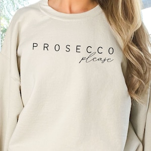 May include: A sand-colored crewneck sweatshirt featuring the black text PROSECCO please printed on the chest. The garment has long sleeves and a relaxed fit. A circular logo with the text Little Swan Crafts is visible in the upper left corner.