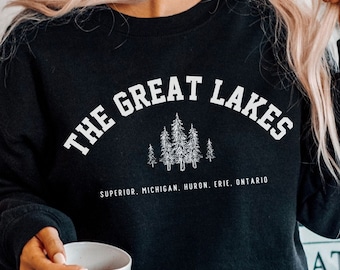 May include: A black crewneck sweatshirt featuring white text that reads THE GREAT LAKES with a graphic of pine trees and the names of the lakes: SUPERIOR, MICHIGAN, HURON, ERIE, ONTARIO. The sweatshirt has long sleeves and a ribbed neckline.