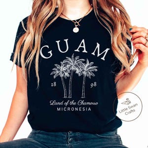 May include: A black short-sleeved t-shirt featuring white text that reads GUAM, 1898, Land of the Chamorro, and MICRONESIA, along with a white line-art illustration of three palm trees centered on the front of the garment.