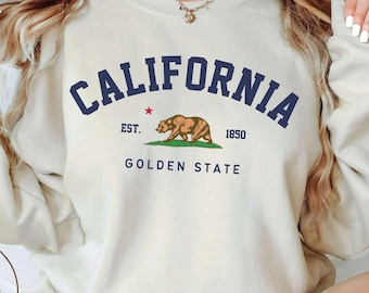 Peut inclure: Un sweat-shirt à col rond couleur sable avec le texte bleu foncé CALIFORNIA arqué au-dessus d'un graphique représentant un ours brun, une petite étoile rouge, le texte EST. 1850 et le texte GOLDEN STATE en lettres bleu foncé.