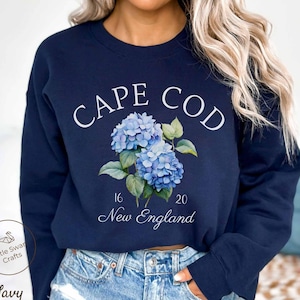 May include: A navy blue crewneck sweatshirt featuring a white graphic print that reads CAPE COD at the top, with a cluster of blue hydrangea flowers in the center, and the text 16 20 New England printed below the floral design.