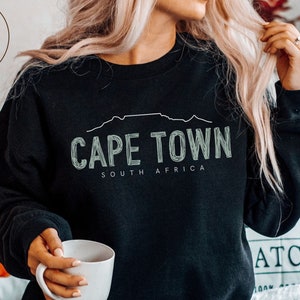 May include: A black long-sleeve crewneck sweatshirt featuring a white line drawing of Table Mountain above the text CAPE TOWN and SOUTH AFRICA in a distressed, textured font. The garment has a ribbed collar, cuffs, and hemline.