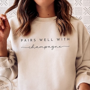 May include: A beige sweatshirt with the text "Pairs well with champagne" in black lettering.